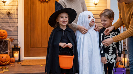 Fototapeta premium Children in witch, ghost and skeleton costumes get ready for trick-or-treating with adults on a decorated Halloween porch, holding colorful candy buckets