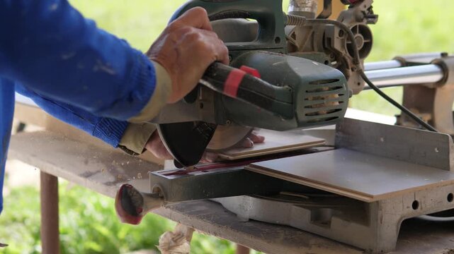 Worker hands rest and grip wood boards beside powered miter saw on outdoor workbench, with blue sleeve, dusty metal housing, bright soft daylight, blurred green background, and