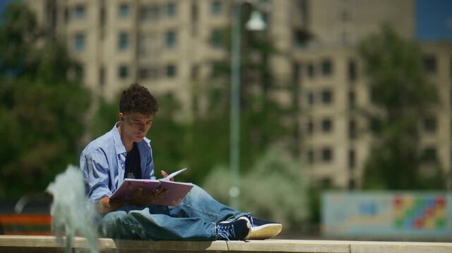 A young man reads a notebook outdoors in a city park, sitting next to a fountain. He is intently focused on his studies in a bright, relaxed urban setting.