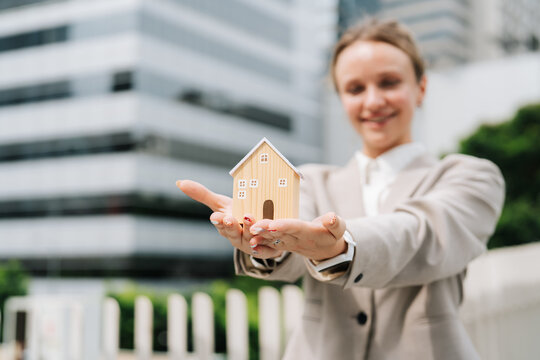 Businesswoman holding small wooden house model on her palm in front of a modern glass office building, real estate investment, property and home ownership