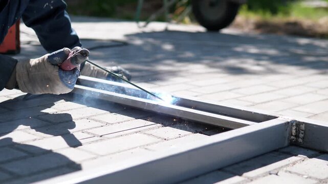 Gloved worker holds welding tool against metal rail on paved outdoor surface, sending bright sparks across silver bars during careful repair work in bright daylight, with welding, worker, gloved