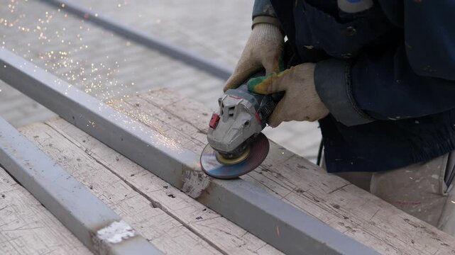 Worker grinding metal, in construction site, with metal beams, safety gloves, grinder, from a close-up slightly elevated angle perspective, with gray, blue accents, and focused and industrious mood.