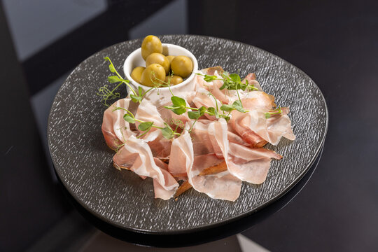 Prosciutto ham served with green olives and microgreens on dark plate