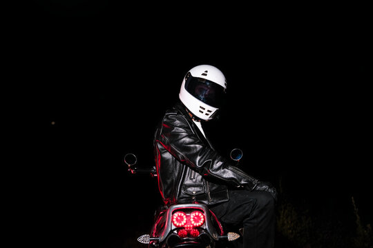 Low key portrait of motorcyclist in white helmet and black leather jacket sitting on motorcycle in dark night. Night ride, dramatic lighting and biker lifestyle concept