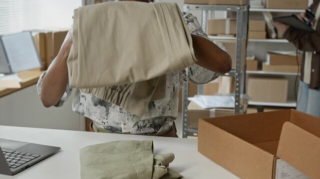 African American young adult woman folding trousers and packing them into cardboard box, camera tilting down. Footage is perfect for e-commerce business promos and retail logistics campaigns
