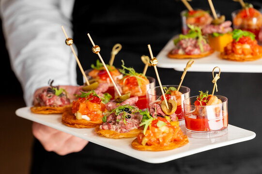 A waiter carries a white tray of elegant gourmet appetizers, featuring seafood tartare, crackers, and shrimp shooters on gold skewers at a catered event.