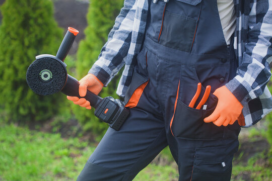 person holding angle grinder outdoors wearing orange gloves and black jacket for home improvement project in garden setting close-up practical and serene mood natural light clean background.