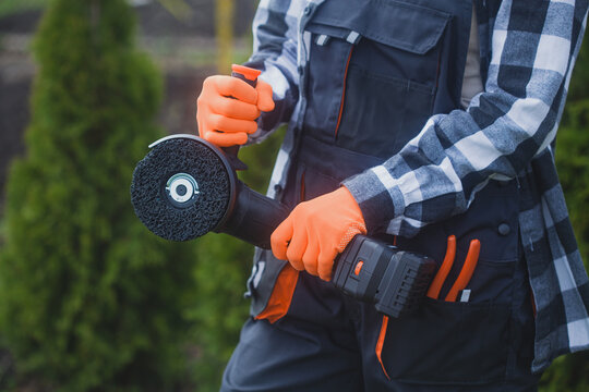 person holding angle grinder outdoors wearing orange gloves and black jacket for home improvement project in garden setting close-up practical and serene mood natural light clean background.