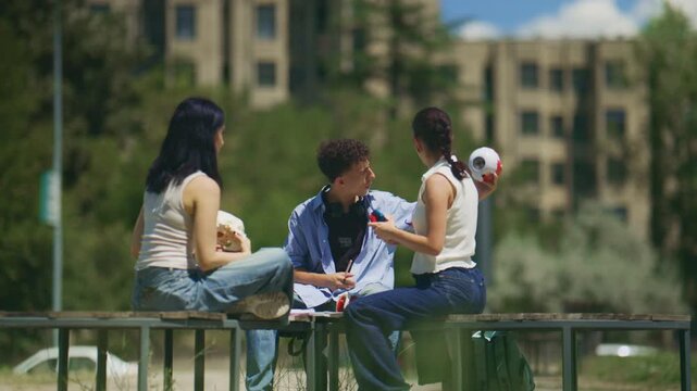 Three young school friends are engaged in a lively discussion while sitting on a bench outdoors. The university students are collaborating and sharing ideas under the bright sun.