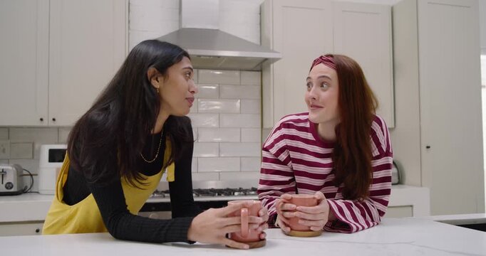Diverse friends leaning in kitchen holding mugs, left friend starting story, right friend sipping
