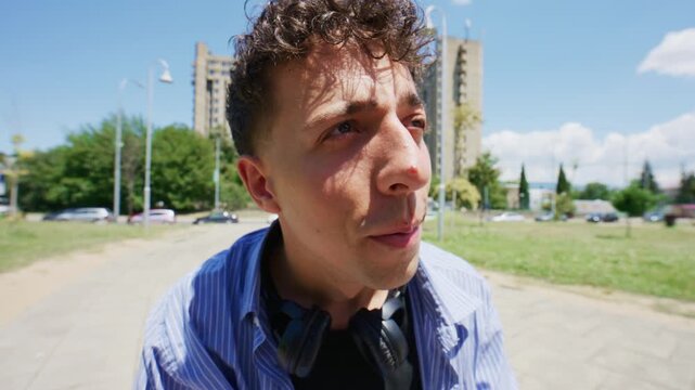 A young man with curly hair plays with a colorful fidget spinner, looking through it like a lens. He seems curious and engaged with the spinning toy in an urban park setting.