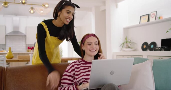 Diverse women sitting on brown couch, friend arriving left pointing at silver laptop sharing joy