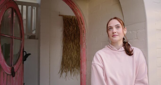 Woman standing entryway by red door, pink hoodie, touching gold earring, smiling, camera tightening