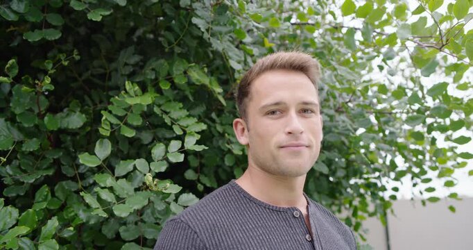 Man standing beside dense shrub turning head and smiling while camera tightening on henley shirt