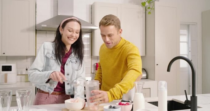 Diverse couple laughing, scooping ice cream into blender cup using scoop, making shake in kitchen