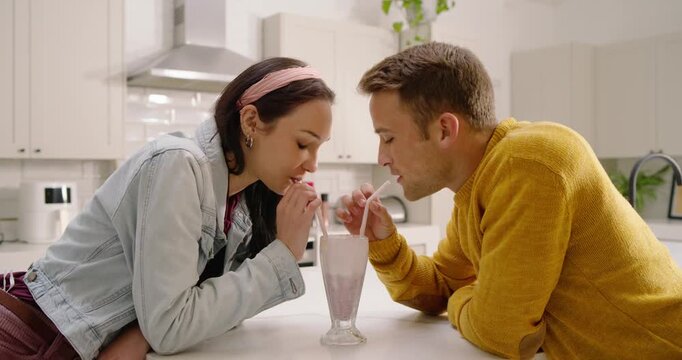 Couple exchanging playful glances, leaning forward and sipping shared milkshake at kitchen counter