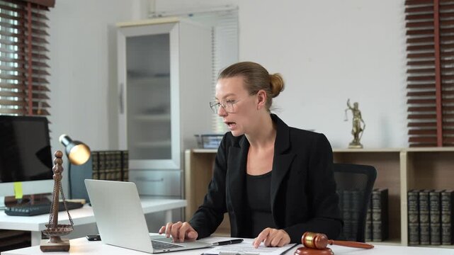 Professional female lawyer providing legal advice and consultation to a client via video conference call using a laptop, sitting at her desk in a modern law firm office