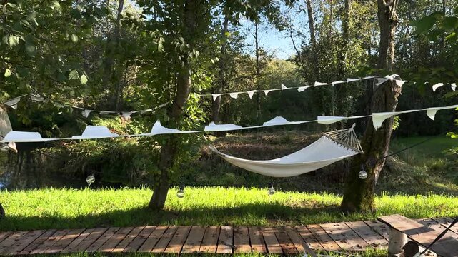 White hammock between trees with garland of white flags next to wooden path during light wind surrounded by lush greenery in summer in sunlight. Relaxation and unity with nature during serene rest