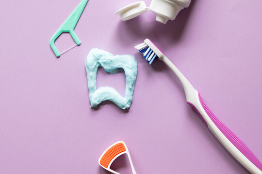 Tooth shape drawn with toothpaste on purple background with dental hygiene tools, toothbrush, tongue scraper, and dental floss.