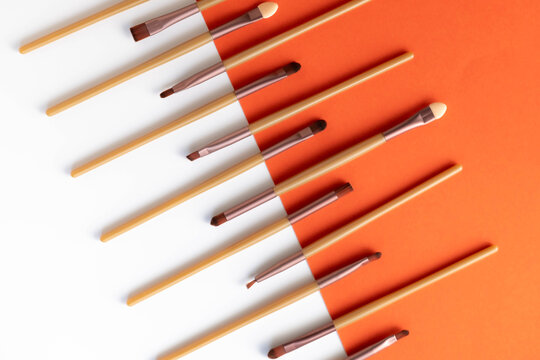 Various sizes and shapes of cosmetic makeup brushes on a bright orange and white background.