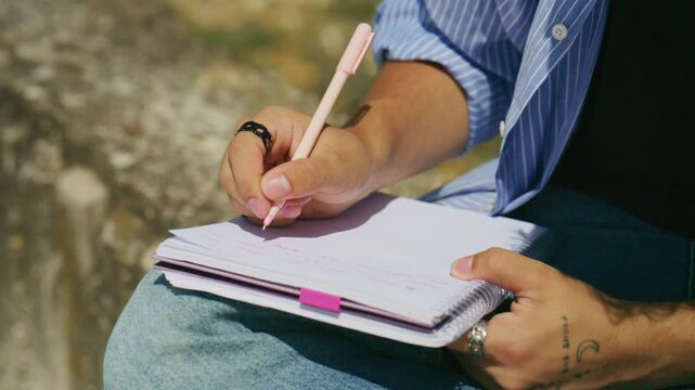 A person's hand meticulously writes in a notebook with a pink pen, capturing thoughts and ideas. The individual is engrossed in their work, making notes on paper in an outdoor setting.