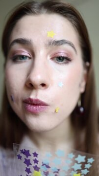 Vertical screen closeup of woman applying colorful star shaped acne patches to clean facial skin during playful skincare and beauty routine in soft daylight at home daily
