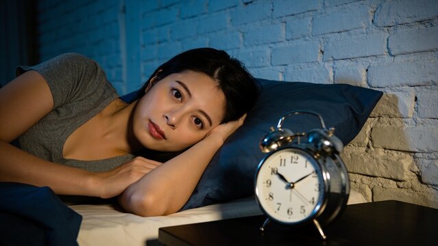 Young Woman Relaxing Awake in Bed at Night with Blue Bedding and Classic Alarm Clock Representing Restful Sleep and Nighttime Routine