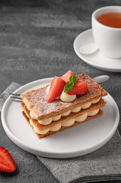 French dessert millefeuille with vanilla cream and fresh strawberries on a white plate on a dark concrete background.