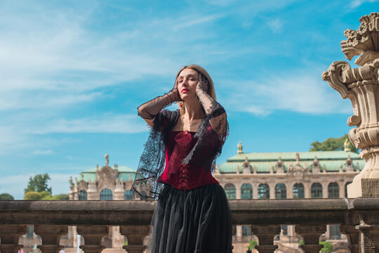 Woman in elegant deep red velvet corset top and black lace cape posing at a historic palace courtyard, fashion and premium lifestyle concept with modern vintage aesthetic