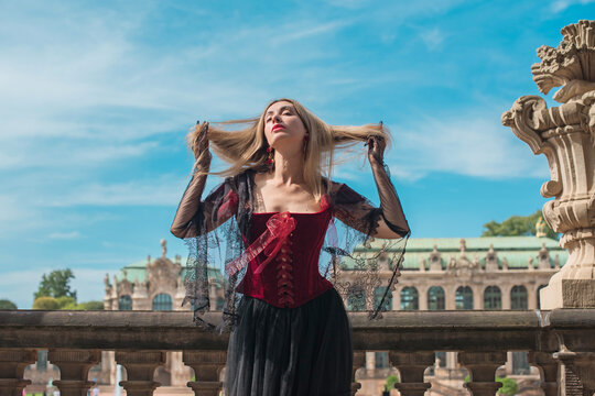 Woman in elegant deep red velvet corset top and black lace cape posing at a historic palace courtyard, fashion and premium lifestyle concept with modern vintage aesthetic