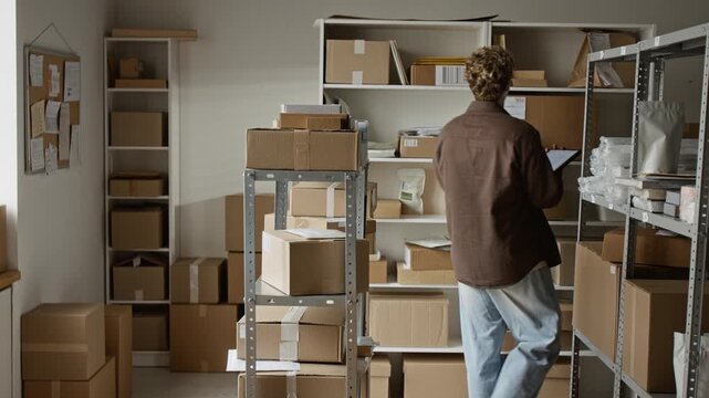 Caucasian young adult man using tablet to manage inventory in small warehouse, checking cardboard boxes on shelves. Clip is ideal for e-commerce, logistics, and small business shipping concepts