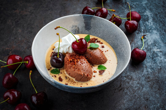 Modern summer dessert - dark black chocolate mousse au chocolat in vanilla sauce with black sweet cherries and meringues served as close-up in bowl - real photo no ai