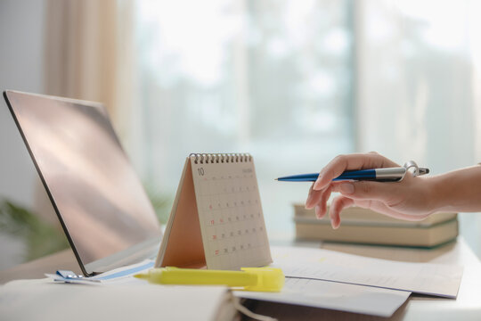 Close up hand holding pen pointing at desk calendar with laptop and papers on table, concept of project planning, schedule management, time management at home and deadline tracking.