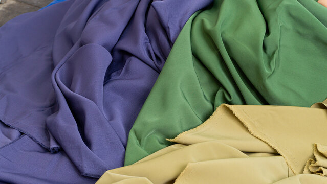 Vivid combination of folded textiles showcasing deep purple, lush green, and warm khaki fabrics draped in overlapping layers. The soft and smooth materials feature graceful waves,