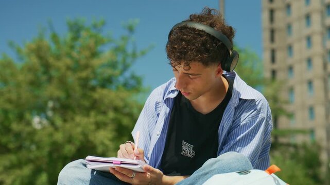 A young man wearing headphones diligently writes in his notebook outdoors. He is focused on his studies or personal thoughts in the park.