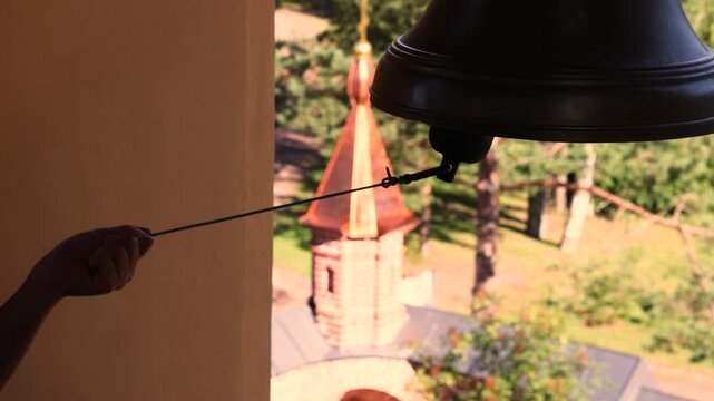 Hand pulling rope to ring a large bronze church bell on a bell tower, blurred orthodox tent roof spire and green treetops far below, sunny daytime elevated religious ceremony scenic outdoor footage