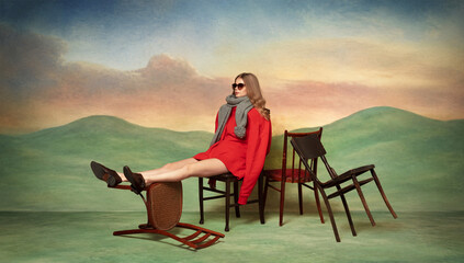 Fototapeta premium Woman in vivid red coat poses among scattered chairs across softly painted theatrical dream landscape. Concept of luxury lookbooks, magazine covers and fashion ads.