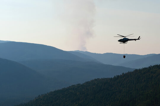A helicopter carries a water bucket to combat a wildfire in mountainous terrain, highlighting the fierce battle against flames and the cooperation between humans and nature.