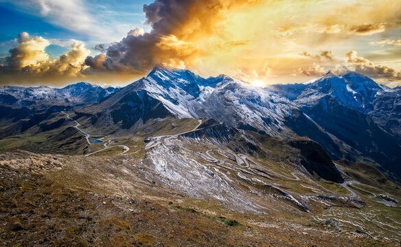 Peaceful picturesque mesmerizing atmospheric landscape with high snow-capped mountains, Gro&szlig;glockner alpine serpentine pass road, and meadow grasslands pasture in sunset