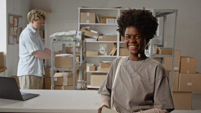 Portrait of smiling African American young adult woman standing in e-commerce warehouse while Caucasian colleague packing boxes. Footage ideal for small business and logistics marketing