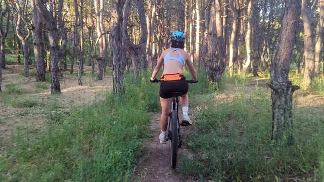 Rear view of a young woman riding a mountain bike along a narrow forest trail, wearing a cycling helmet and athletic sportswear, surrounded by trees and lush green vegetation