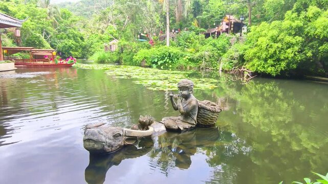 sculpture of boy playing flute while riding buffalo in pond. park sculpture showing rural life scene with water buffalo. concept of sculpture art and cultural heritage in tropical park