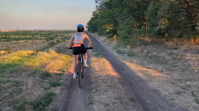 Rear view of a young woman riding a mountain bike on a dirt trail between a harvested field and trees at golden hour, with a distant city skyline in the background