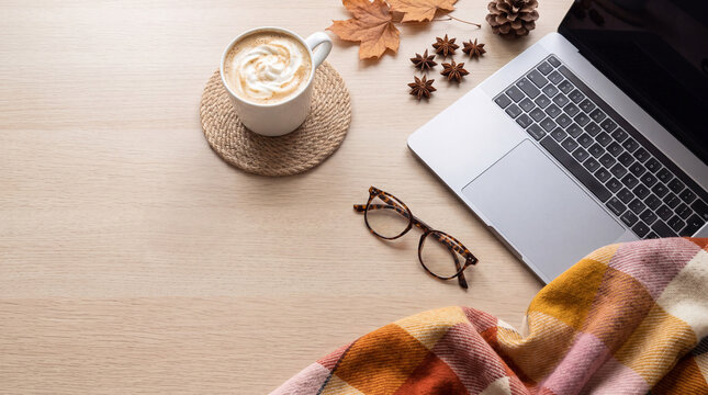 A cozy autumn workspace flat lay featuring a laptop, a cup of coffee, eyeglasses, fall leaves, and a plaid blanket on a wooden table, perfect for seasonal concepts.