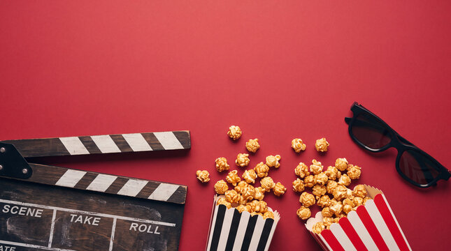 Flat lay of movie night items including a clapperboard, spilled popcorn in striped boxes, and 3D glasses on a red background with copy space.