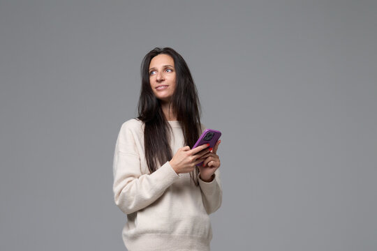 Woman with smartphone looking away with thoughtful expression on grey background