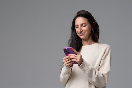 Smiling woman using smartphone with purple case looking down on grey background