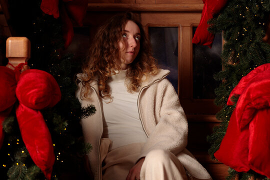 A young woman with curly hair makes a humorous grimace while sitting indoors amidst festive Christmas greenery and red bows