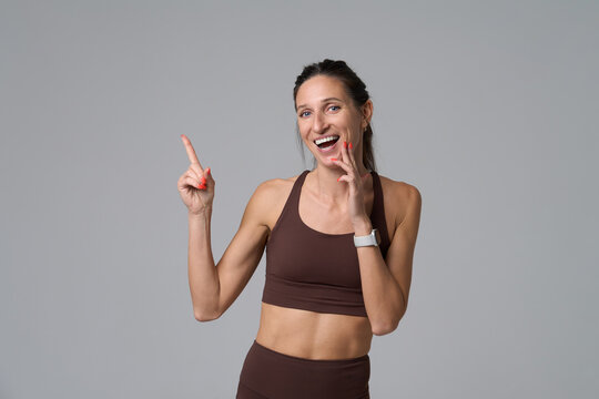 Cheerful woman in brown sportswear pointing finger at copy space on gray background
