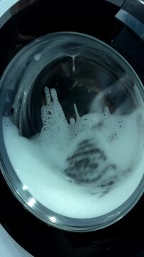 Close up shot of laundry rotating inside a modern washing machine with white soap foam and water bubbles.
